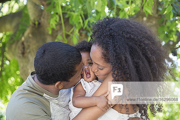 Liebevolle Eltern küssen Kleinkind-Tochter unter Baum