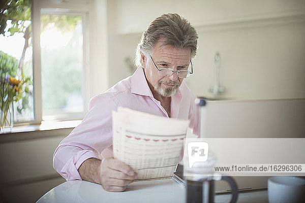 Älterer Mann liest Zeitung am Laptop am morgendlichen Küchentisch