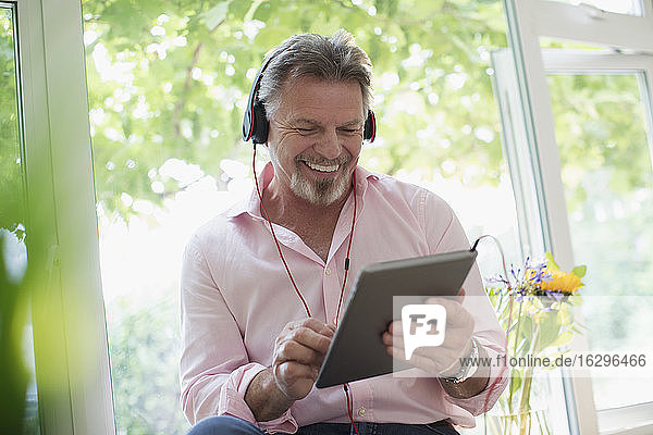 Glücklicher älterer Mann mit Kopfhörer und digitalem Tablet am Fenster