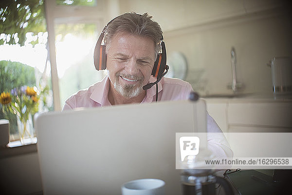 Glücklicher älterer Mann mit Kopfhörern arbeitet am Laptop in der Morgenküche