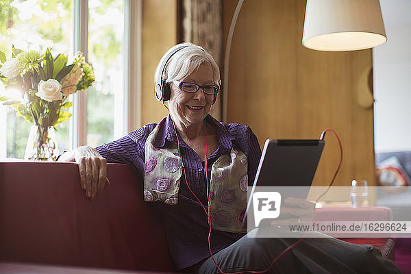 Lächelnde ältere Frau mit Kopfhörer und digitalem Tablett auf dem Sofa