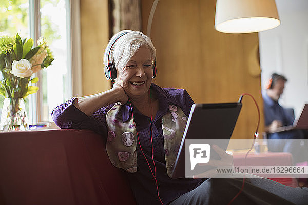 Glückliche ältere Frau benutzt Kopfhörer und digitales Tablett auf dem Sofa