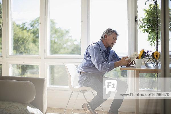 Älterer Mann liest Buch am Esstisch am sonnigen Fenster
