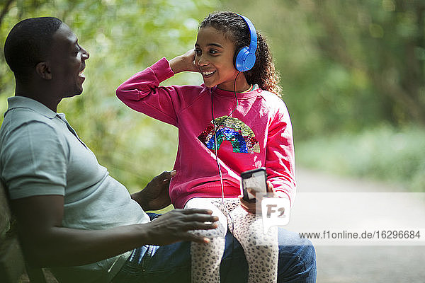 Happy father and daughter sitting on park bench with headphones