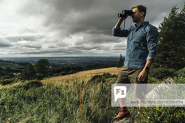 Männlicher Wanderer  der durch ein Fernglas schaut  während er in der Landschaft vor einem bewölkten Himmel steht