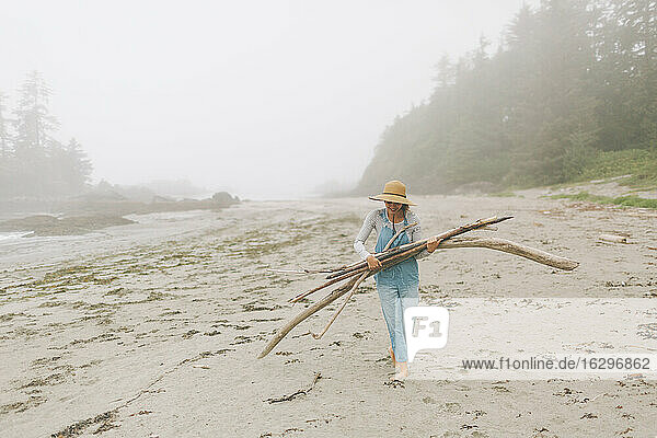 Frau mit Hut trägt Baumstämme beim Spaziergang am Strand