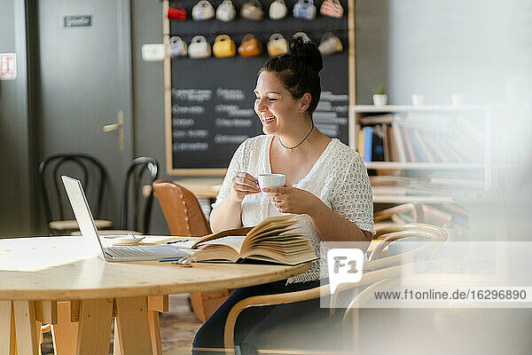 Smiling young woman holding coffee cup while looking at laptop on table in cafe