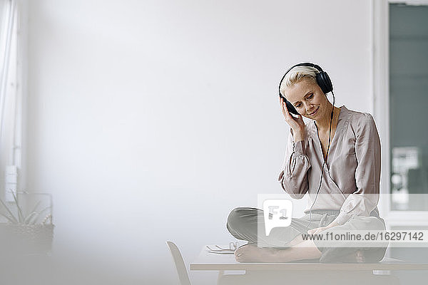 Weibliche Fachkraft hört Musik über Kopfhörer  während sie auf dem Schreibtisch an der Wand im Büro sitzt