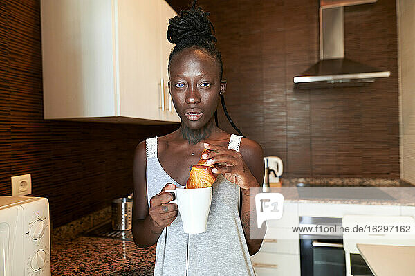 Junge Frau isst Croissant mit Kaffee in der Küche zu Hause