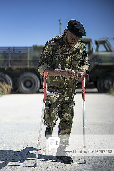 Injured army soldier with crutches text messaging on smart phone during sunny day