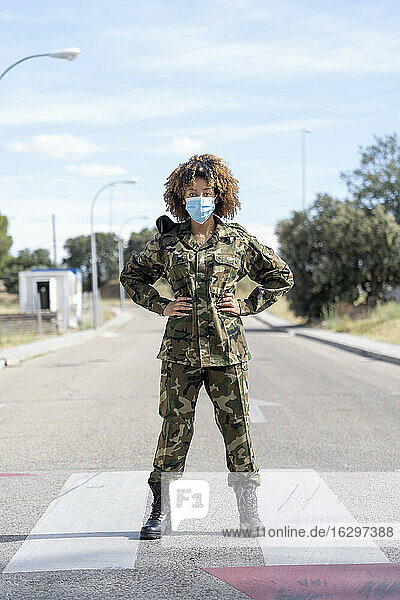 Weiblicher Armeesoldat mit Schutzmaske und Händen auf der Hüfte steht auf der Straße