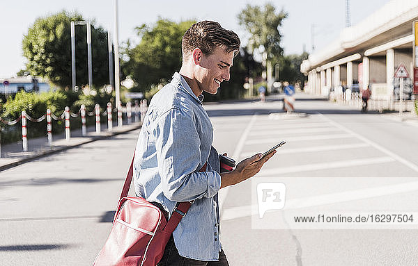 Lächelnder junger Mann  der ein Mobiltelefon benutzt  während er in der Stadt auf der Straße geht