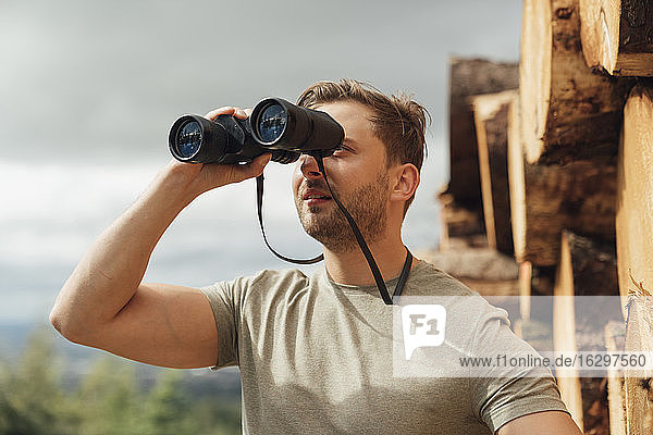 Mid adult man looking through binoculars against sky in forest
