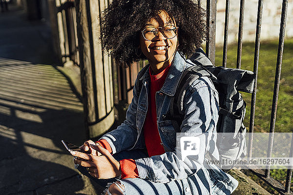 Smiling young woman with afro hair using smart phone while sitting by railing