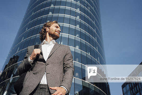 Lächelnde gut aussehende junge männliche Fachkraft schaut weg  während sie vor einem modernen Bürogebäude in der Innenstadt steht