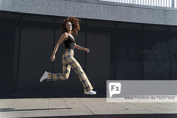 Lächelnde Frau mit zerzaustem Haar  die auf dem Bürgersteig in der Stadt springt