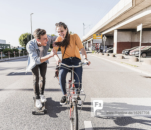 Verspielter Freund auf dem Skateboard  während seine Freundin auf der Straße in der Stadt Fahrrad fährt