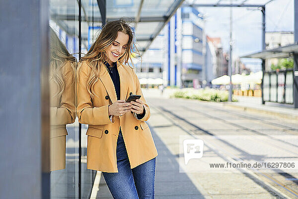 Lächelnde Frau  die ein Smartphone benutzt  während sie an der Straßenbahnhaltestelle wartet