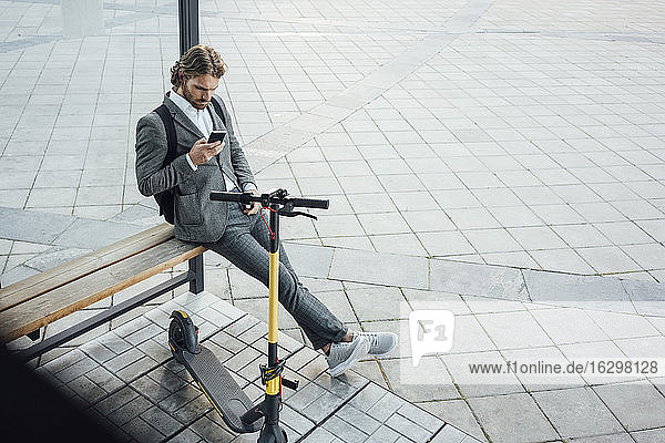 Männlicher Unternehmer  der ein Mobiltelefon benutzt  während er auf einer Bank mit einem Elektroroller in der Innenstadt sitzt