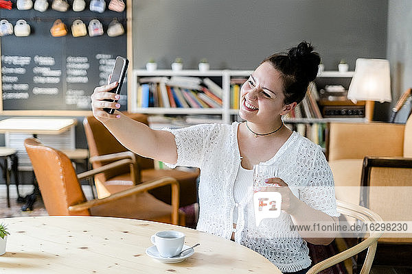 Lächelnde Frau  die ein Getränk hält und ein Selfie mit ihrem Smartphone am Tisch im Café macht