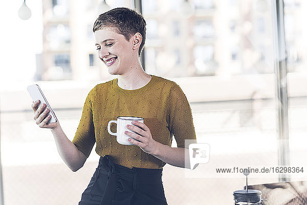 Lächelnde Geschäftsfrau im Büro mit Kaffeetasse und Mobiltelefon