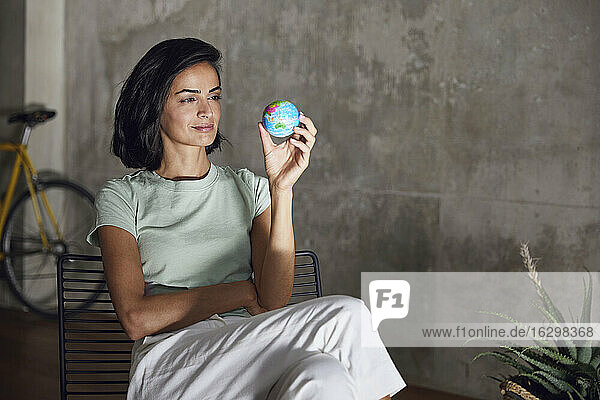 Geschäftsfrau  die einen kleinen Globus hält und auf einem Stuhl an der Wand im Büro sitzt