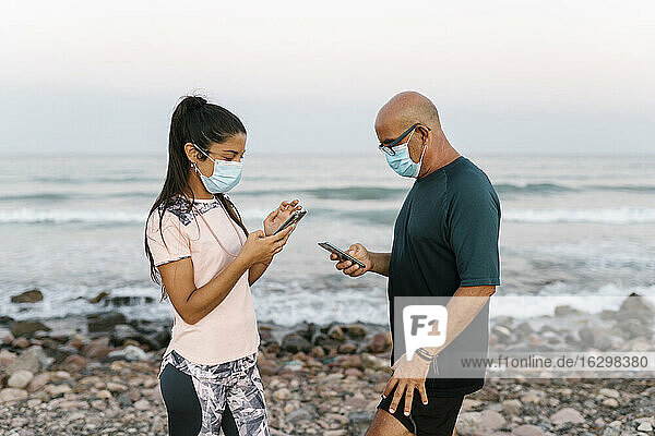 Woman and senior man with face mask using phone while standing against sea
