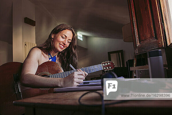 Lächelnde junge Frau  die in ein Buch schreibt  während sie zu Hause Gitarre übt