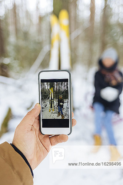 Boyfriend photographing girlfriend by skis through mobile phone in forest during winter