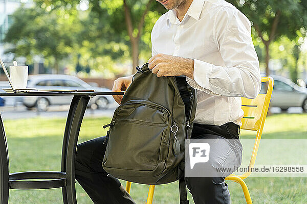 Businessman zipping backpack while sitting on chair at sidewalk cafe