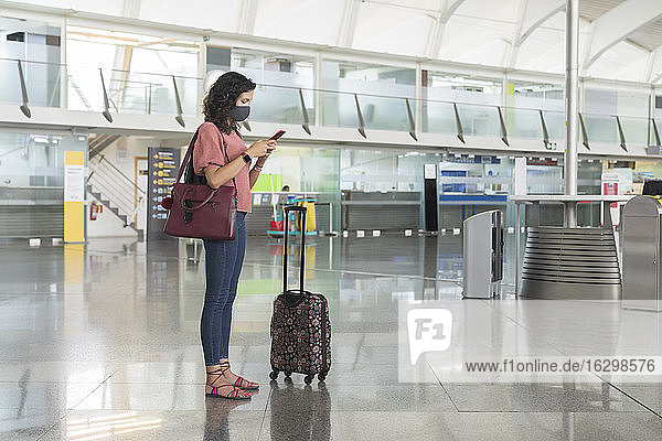 Junge Frau mit Gesichtsschutzmaske schreibt Textnachrichten auf ihrem Smartphone  während sie am Flughafen steht