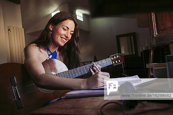 Lächelnde junge Frau  die in ein Buch schreibt  während sie zu Hause Gitarre übt