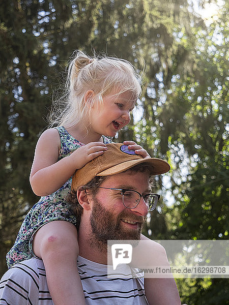 Lächelnder Vater trägt seine Tochter auf der Schulter in einem öffentlichen Park