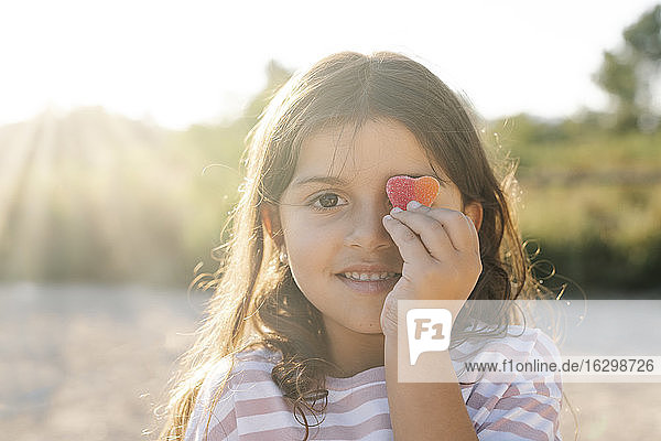 Nahaufnahme von niedlichen lächelnden Mädchen hält Süßigkeiten Herz gegen Gesicht im Park bei Sonnenuntergang