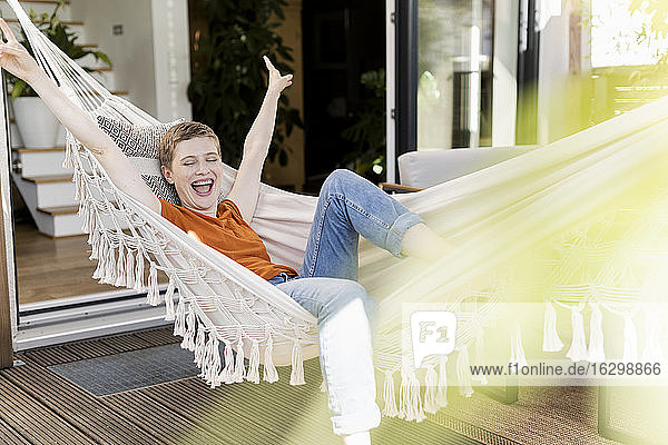 Cheerful woman with arms raised screaming while lying on hammock against house in porch