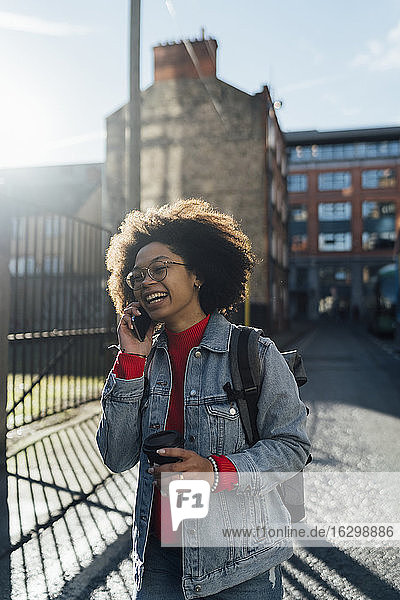 Fröhliche Afro-Frau  die über ein Smartphone spricht  während sie mit einer Kaffeetasse auf der Straße steht