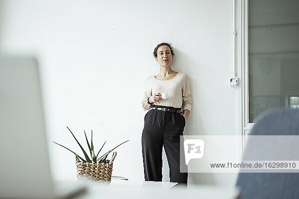 Selbstbewusste Geschäftsfrau mit Hand in der Tasche  die eine Kaffeetasse hält  während sie vor einer weißen Wand steht