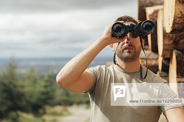 Male hiker looking through binoculars while standing against sky in forest