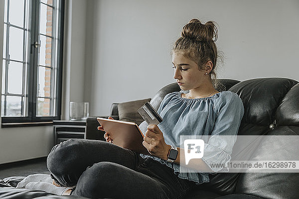 Girl with credit card doing online shopping over digital tablet while sitting on sofa at home