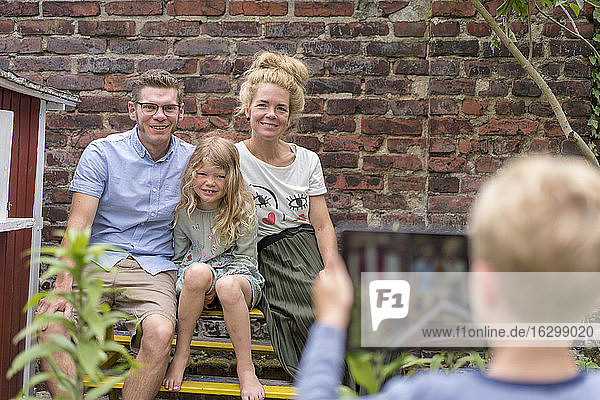 Junge fotografiert Familie sitzt gegen Backsteinmauer durch digitale Tablette im Hinterhof