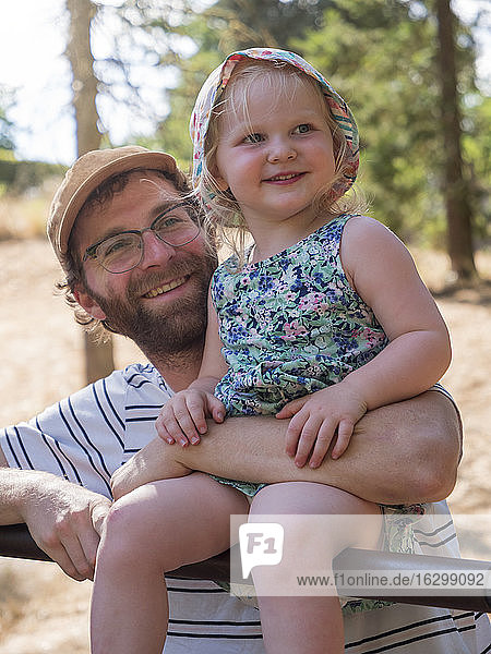 Vater hält seine Tochter auf dem Arm  während er in einem öffentlichen Park auf einer Stange sitzt