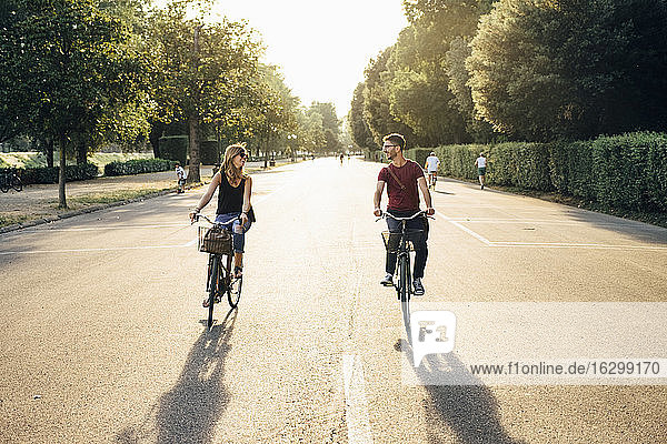Happy couple cycling on road amidst trees in park during sunset