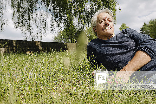 Nachdenklicher Mann mit Tasse auf der Seite liegend über Gras im Hinterhof