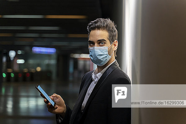 Geschäftsmann mit Maske  der ein Smartphone benutzt  während er an der Wand eines Bahnhofs steht