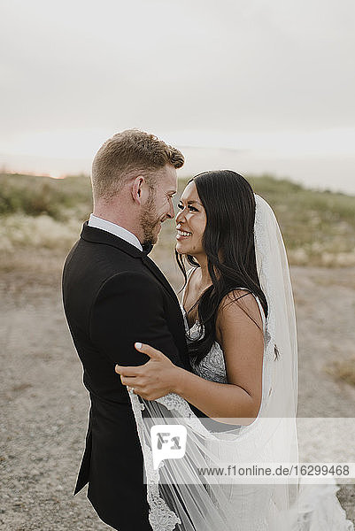 Smiling bridegroom looking at each other in field against sky
