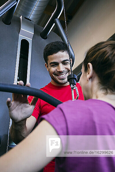 Lächelnder Fitnesstrainer  der einer Frau beim Training mit seitlichen Pull-Down-Gewichten hilft