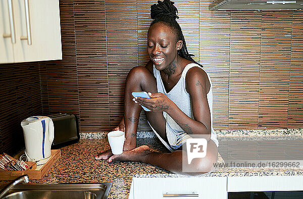Lächelnde Hipster-Frau  die ein Smartphone benutzt  während sie zu Hause auf dem Küchentisch sitzt