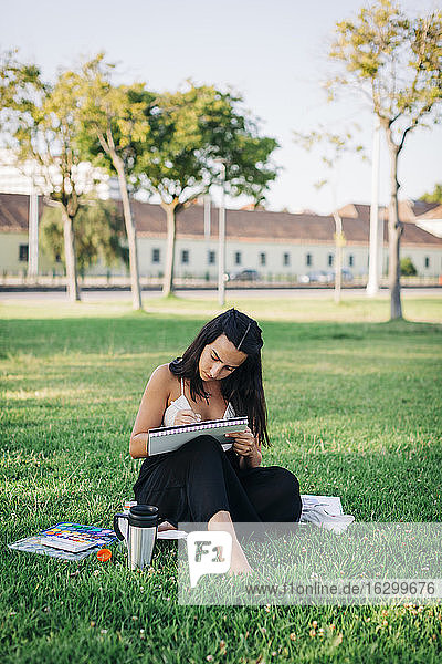 Junge Frau malt mit Aquarellfarbe in einem Buch  während sie in einem öffentlichen Park sitzt