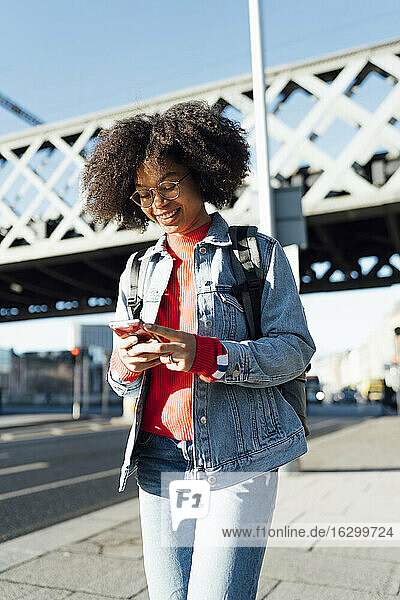 Afro junge Frau  die ein Smartphone benutzt  während sie auf dem Bürgersteig vor einer Brücke in der Stadt steht