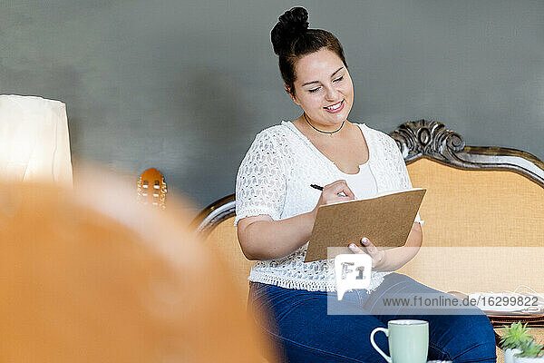 Lächelnde junge Frau schreibt in einem Buch und sitzt auf einem Stuhl in einem Café